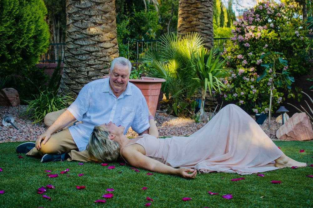 Couple relaxing on lush green garden lawn surrounded by tropical plants, woman in pink dress lying across mans lap.