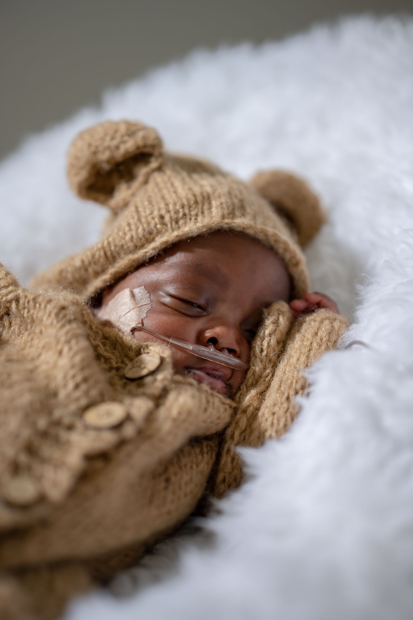 A sleeping baby wears a cozy brown knit bear outfit on white fluffy bedding.