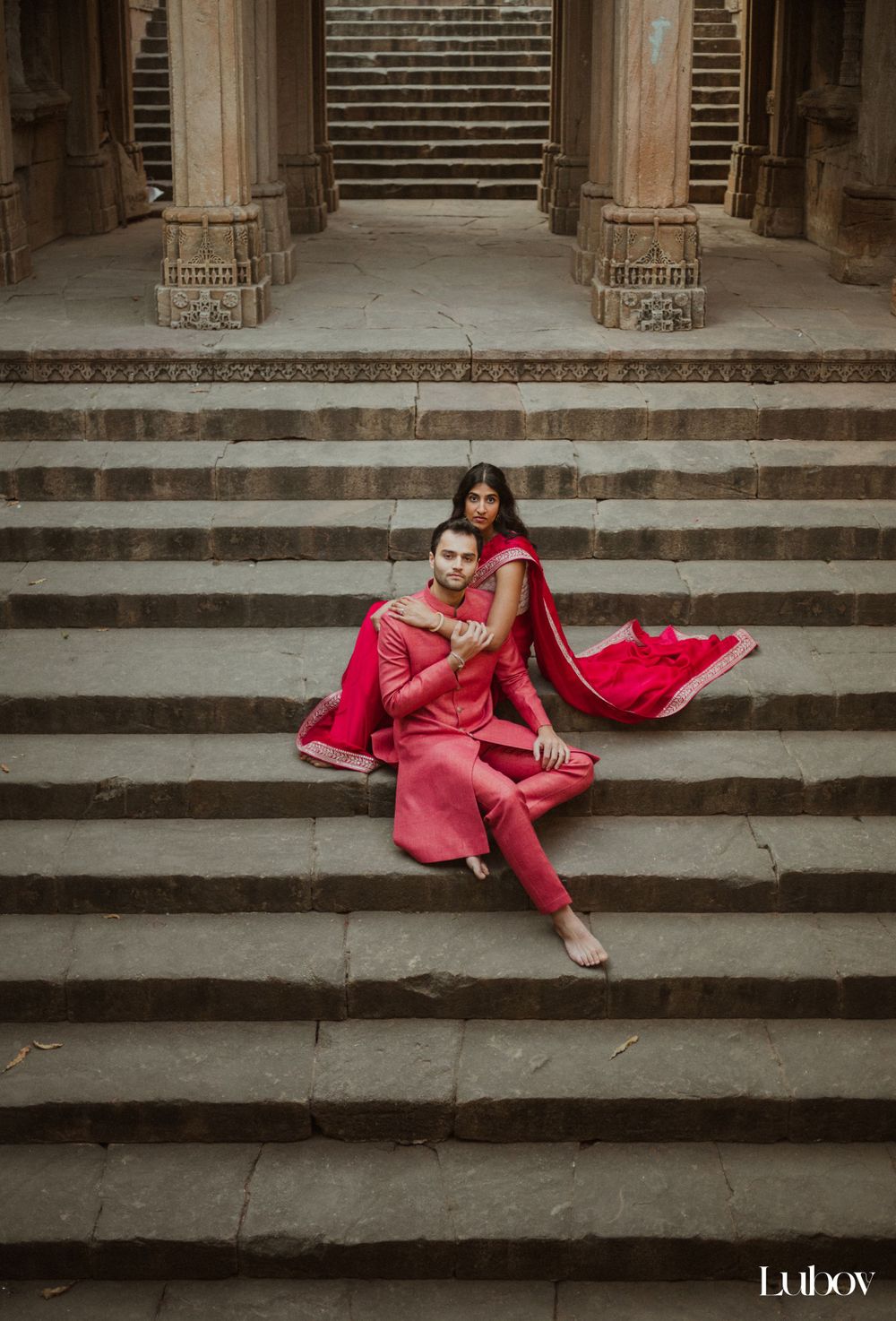 Couple in vibrant red traditional Indian attire seated on stone temple steps.