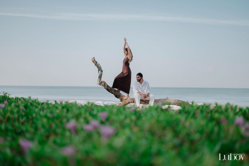 Couple poses together on a beach with waves and green foliage in the background.