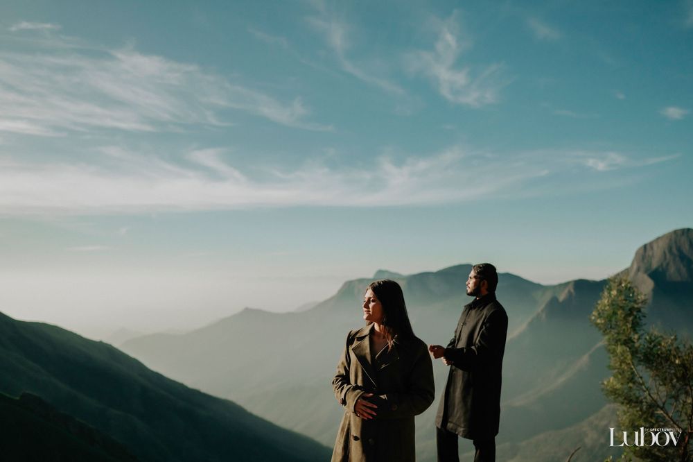 Two silhouettes stand on a mountain overlook during sunset with misty peaks in the distance.