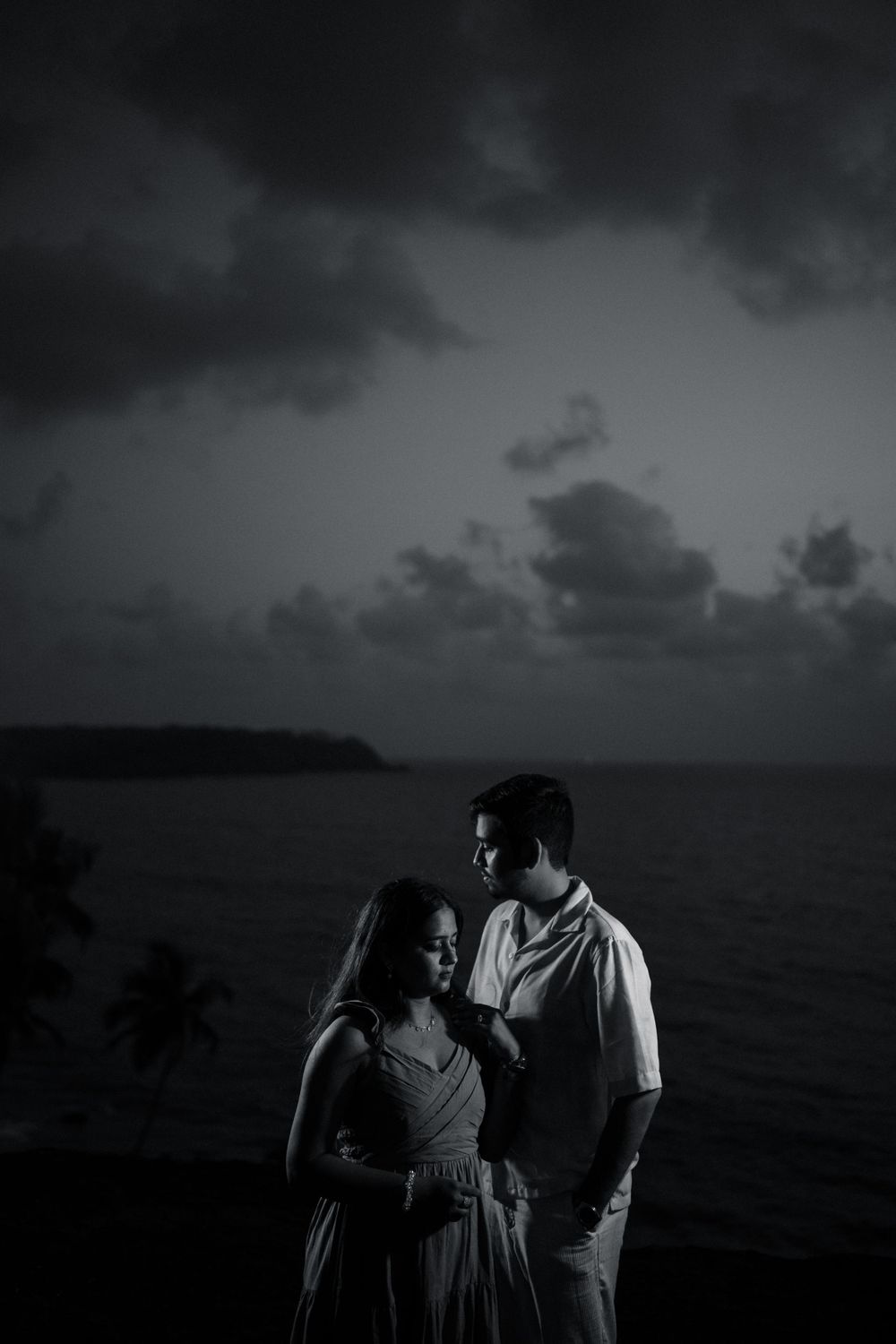 Romantic couple embracing against dramatic stormy ocean sunset in black and white.