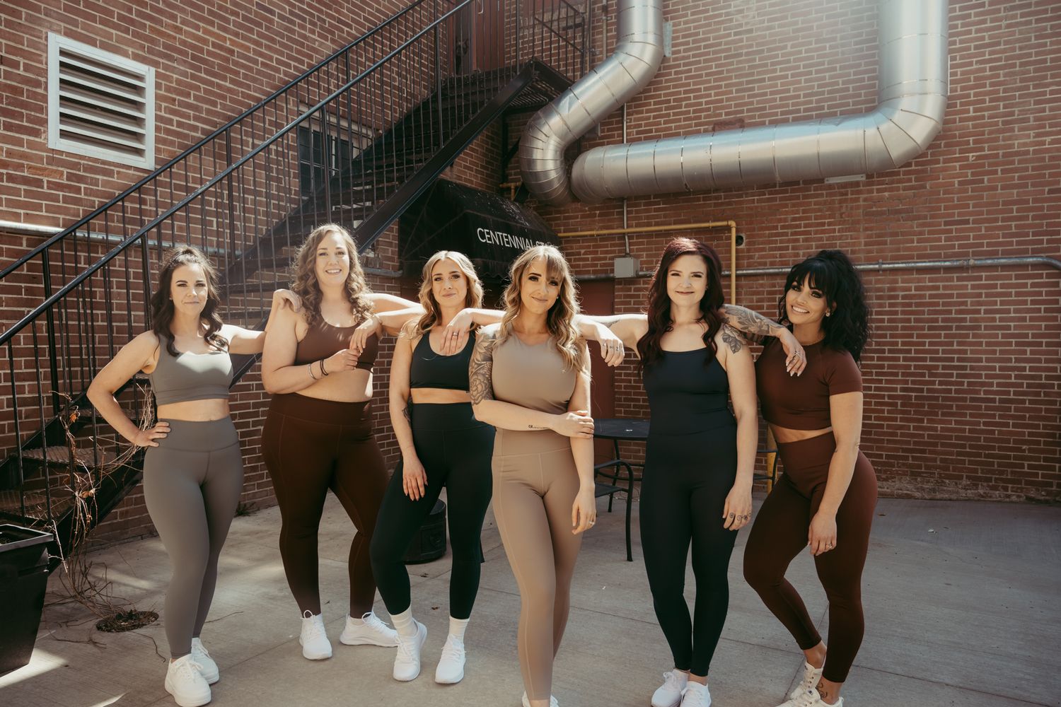 Group of athletic women in workout attire posing together against a brick wall background.