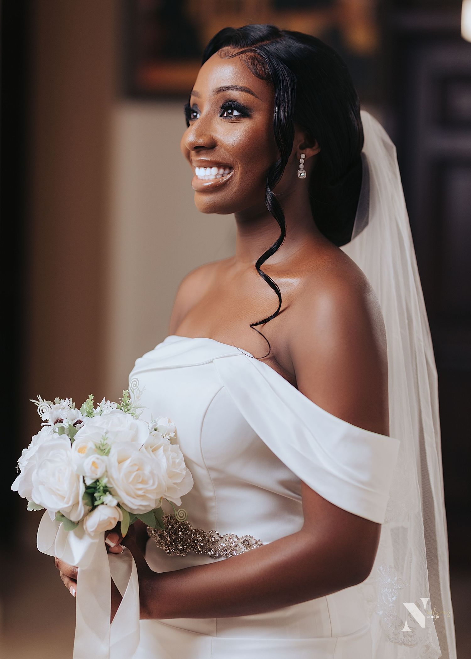 A radiant bride in a white off-shoulder wedding gown holds a bouquet of white roses while wearing a flowing veil.