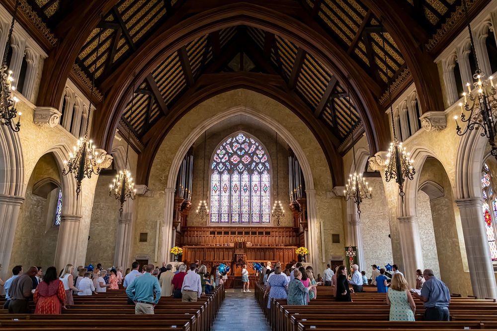 Interior view of a grand cathedral with arched ceilings, stained glass windows, and congregation seated in wooden pews.