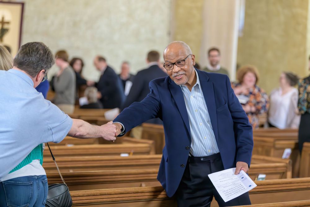 A person in a navy blazer gestures while holding papers during a gathering in a church sanctuary.