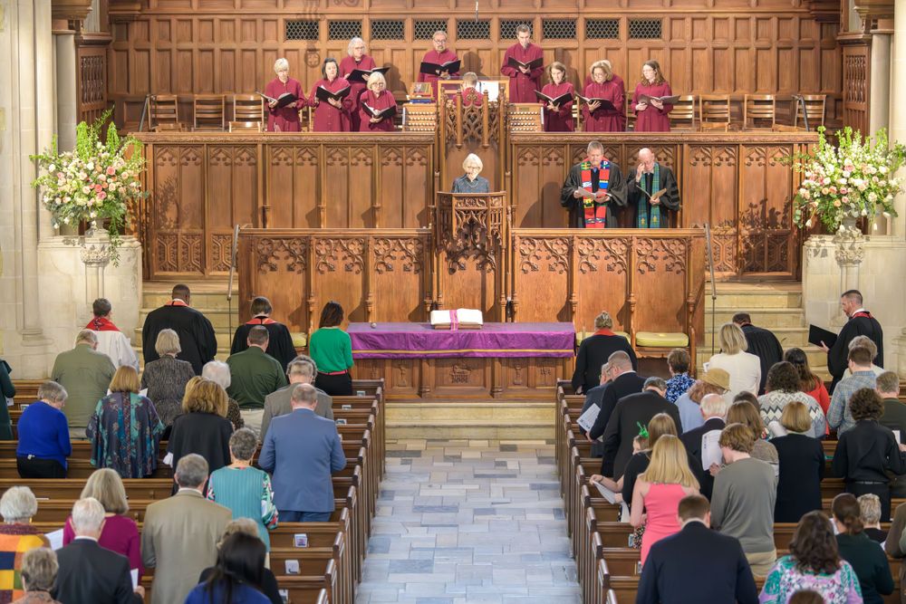 People gathered in a church sanctuary with wooden decor and choir members wearing pink robes in the choir loft.