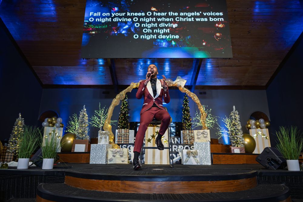 Stage performers in colorful costumes stand on a theatrical set with blue lighting and Christmas trees in the background.