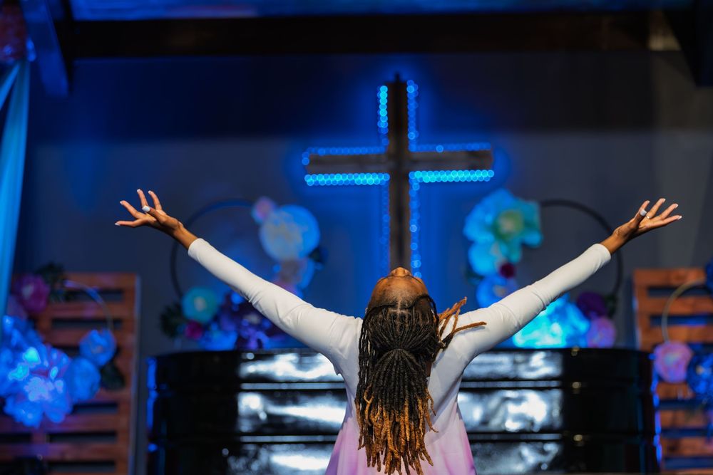 Woman raises arms in worship before a glowing neon cross on a decorated church stage.