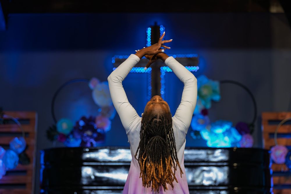 Woman with braids raises arms in worship before illuminated cross in church setting.