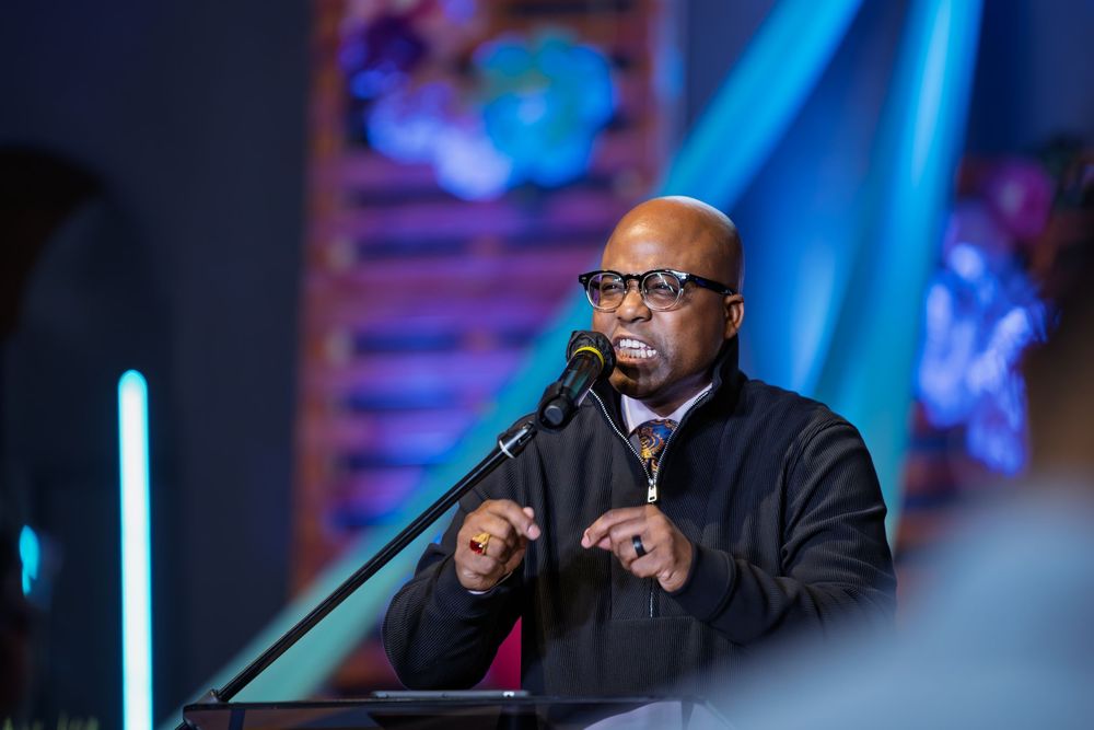 Man in glasses speaking at a podium with a microphone at an evening event with colorful lights.
