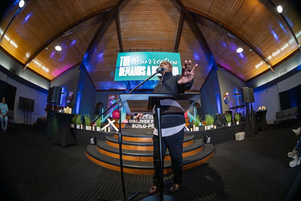 A speaker addresses a congregation from a circular pulpit in a modern church with a large projection screen behind.