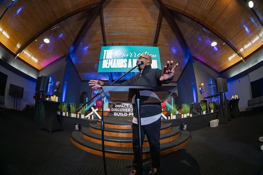 Speaker addresses congregation in a wood-ceiling church with colorful stage lighting and projection screen.