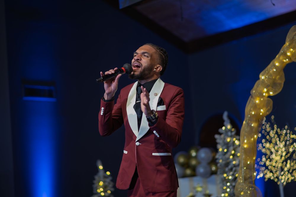 A performer in a burgundy suit jacket sings passionately on stage with dramatic blue lighting and gold decor.