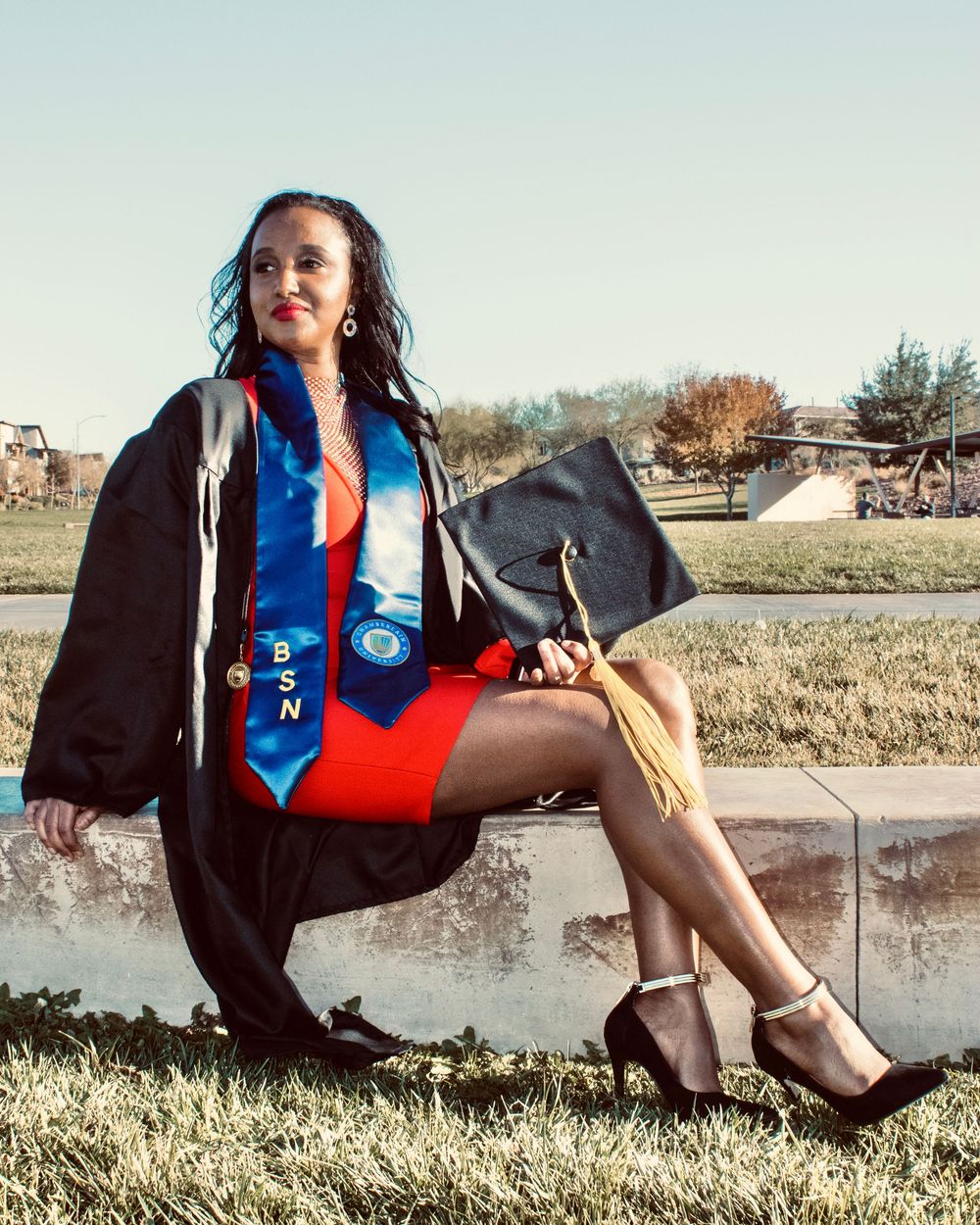 BSN nursing graduate posing confidently in cap and gown while seated on an outdoor ledge.