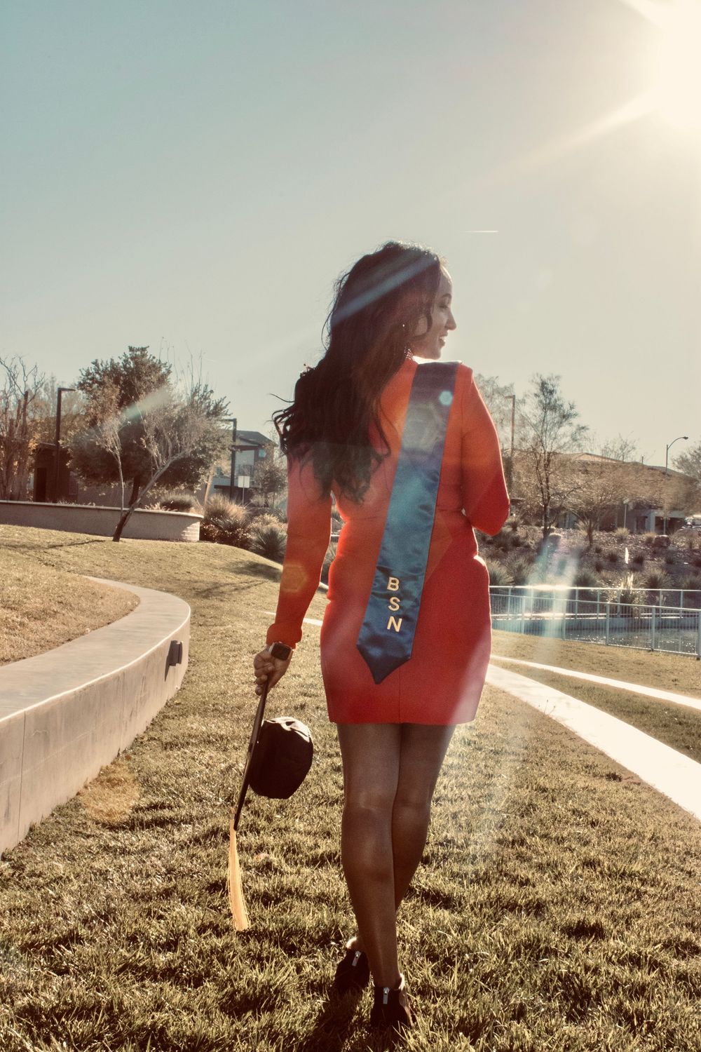 BSN nursing graduate in red dress walking outdoors holding graduation cap in sunlight.