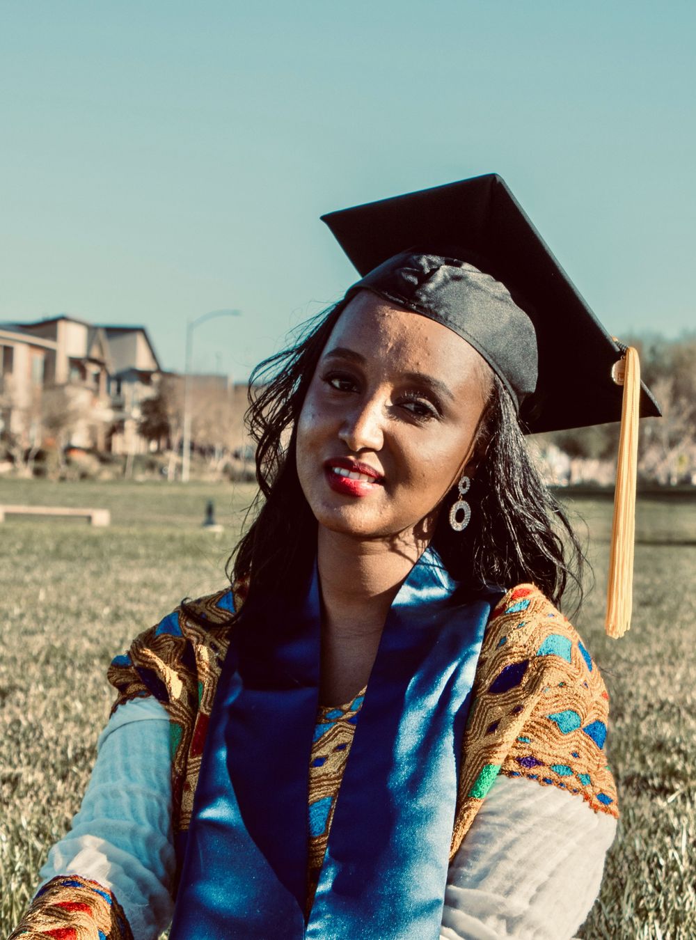 Smiling graduate in cap and colorful cultural attire sitting on grass in an outdoor setting.