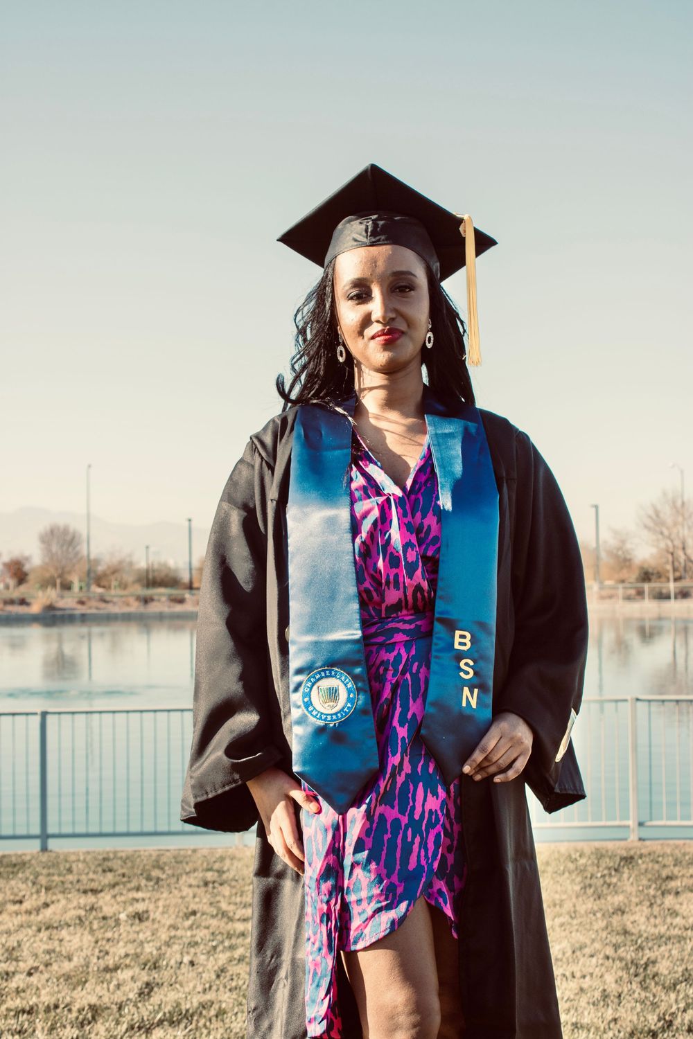 Woman in black graduation gown with BSN stole and pink dress stands near a lake.