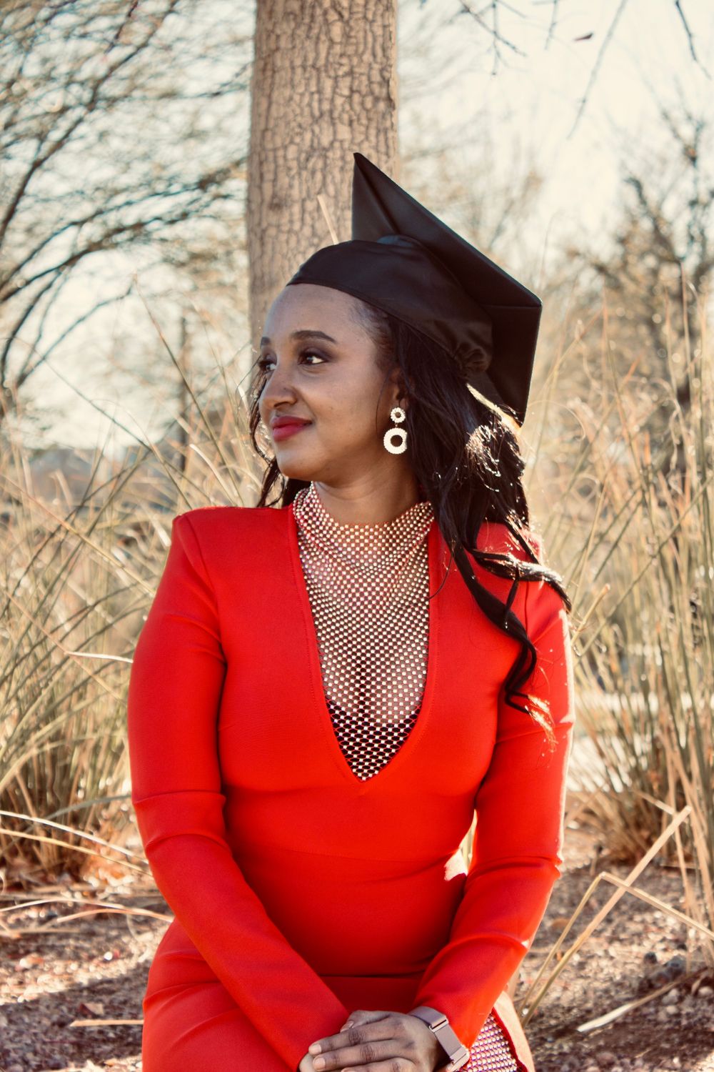 Graduate in red blazer and black mortarboard cap poses outdoors among dry grasses.