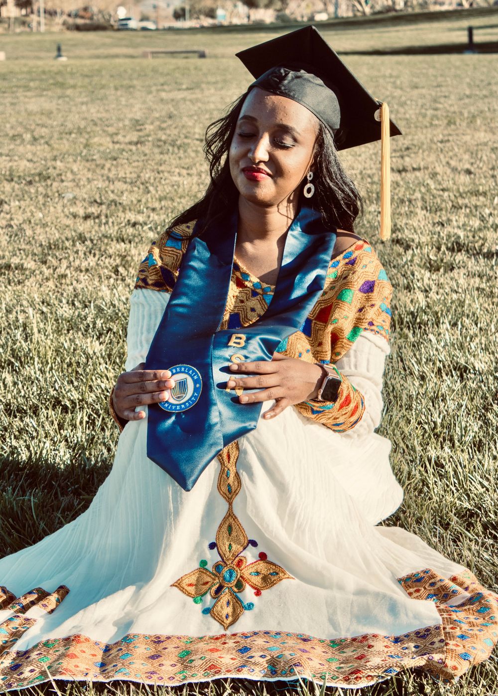 Graduate in white traditional dress with BSN stole and mortarboard sits on grass.