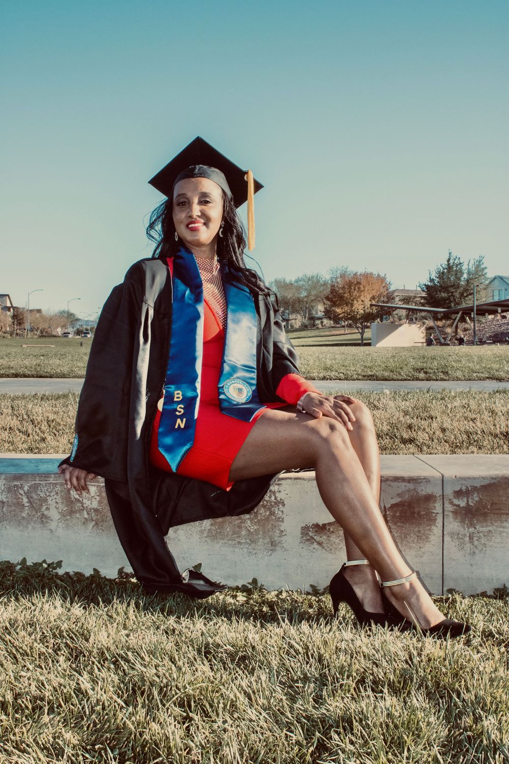 Graduate in black cap and gown with BSN stole sits on concrete wall outdoors in red dress.