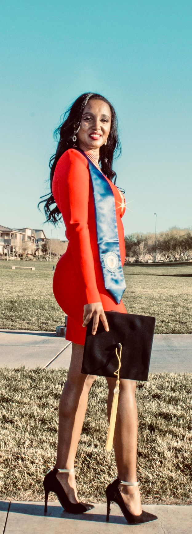 Woman in red dress and blue BSN graduation stole poses outdoors holding mortarboard cap in heels.