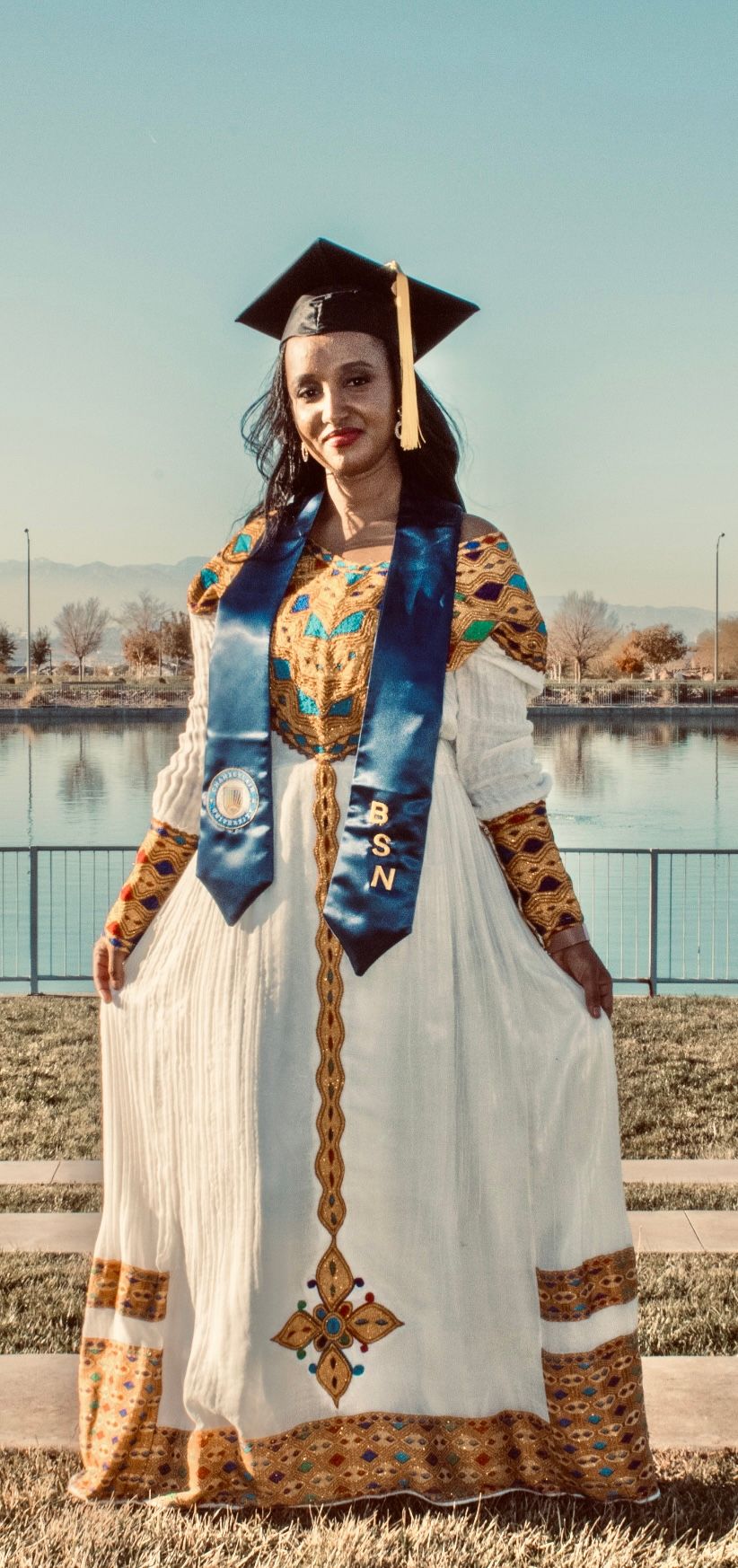 Woman in traditional Ethiopian dress with gold embroidery wears BSN graduation cap and stole outdoors.