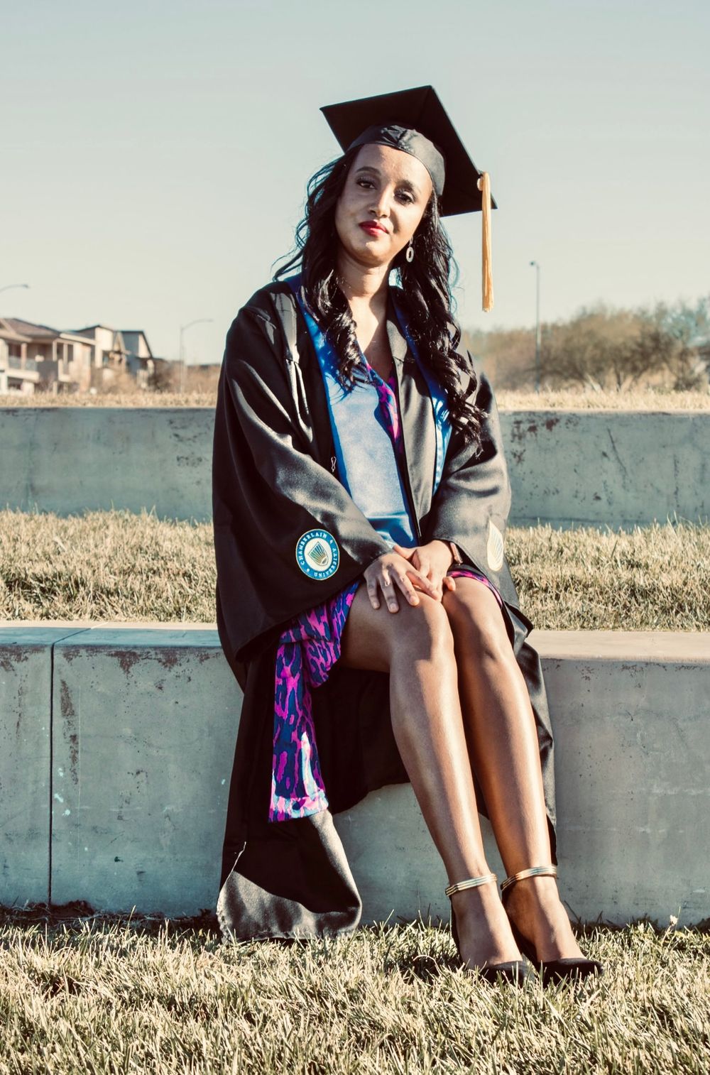 Graduate in black cap and gown with colorful stole sits on concrete ledge outdoors in heels.