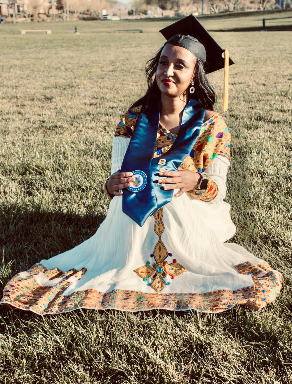 Graduate in traditional white Ethiopian dress sits on grass wearing black mortarboard and blue BSN stole.