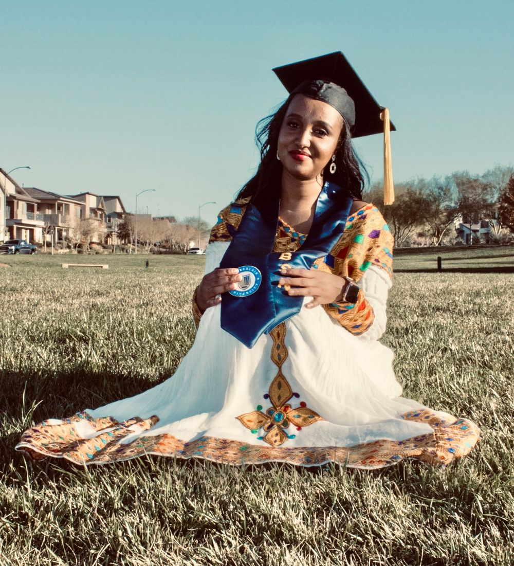 Woman in traditional Ethiopian dress with gold embroidery sits on grass wearing black graduation cap.