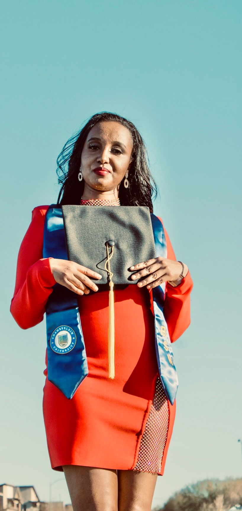 Woman in red dress holds graduation cap while wearing blue BSN stole against clear blue sky.
