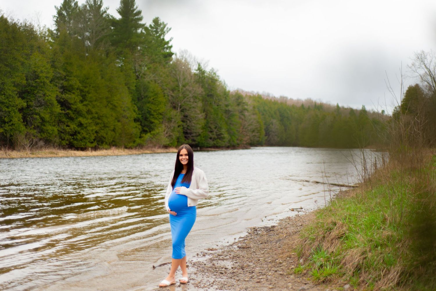 A maternity photoshoot by a scenic river with evergreen trees in the background.