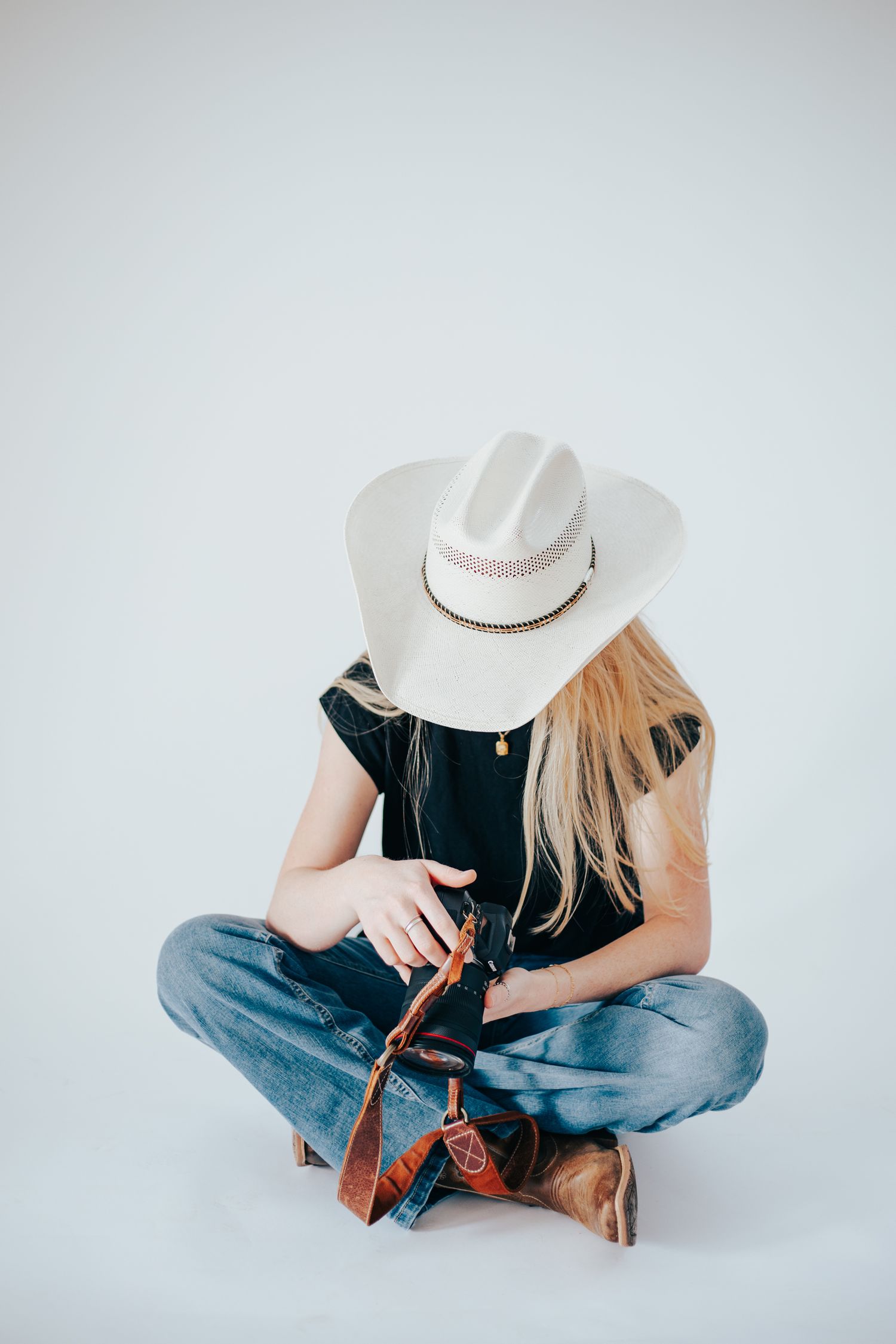 A person in western attire sits cross-legged examining a camera strap against white background.