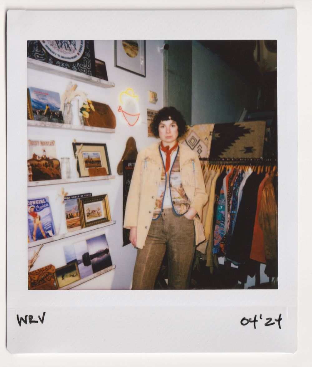 Person wearing tan jacket poses in vintage clothing store with wall display of framed photos and artwork.