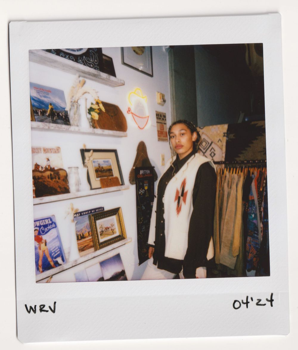 Shopper in black and white outfit poses near photo wall display in vintage clothing store.