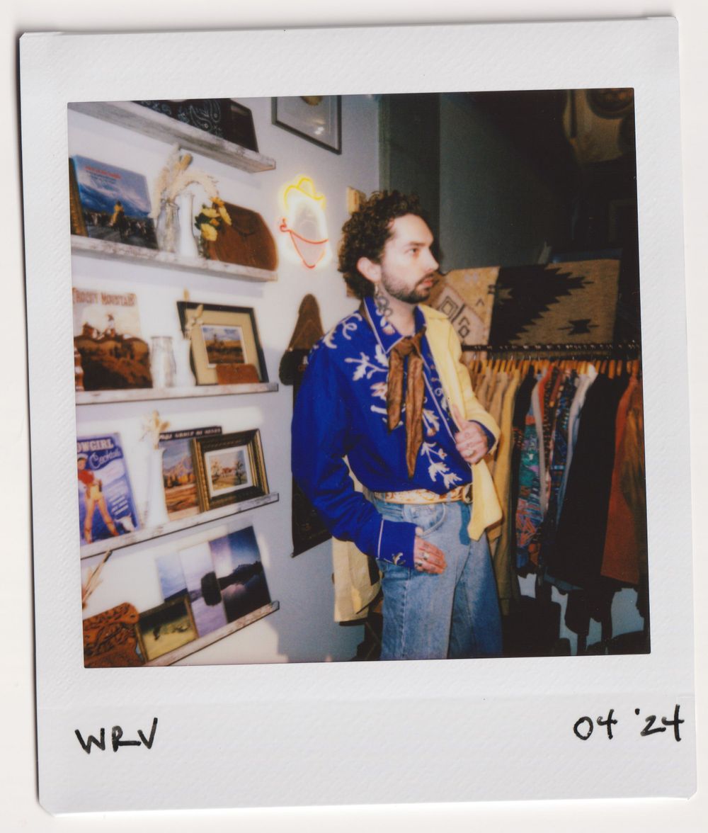 Shopper wearing blue jacket poses near clothing racks and wall art in vintage store.