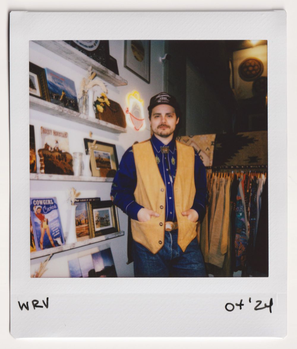 Customer in yellow vest and baseball cap stands near photo wall display in vintage clothing shop.