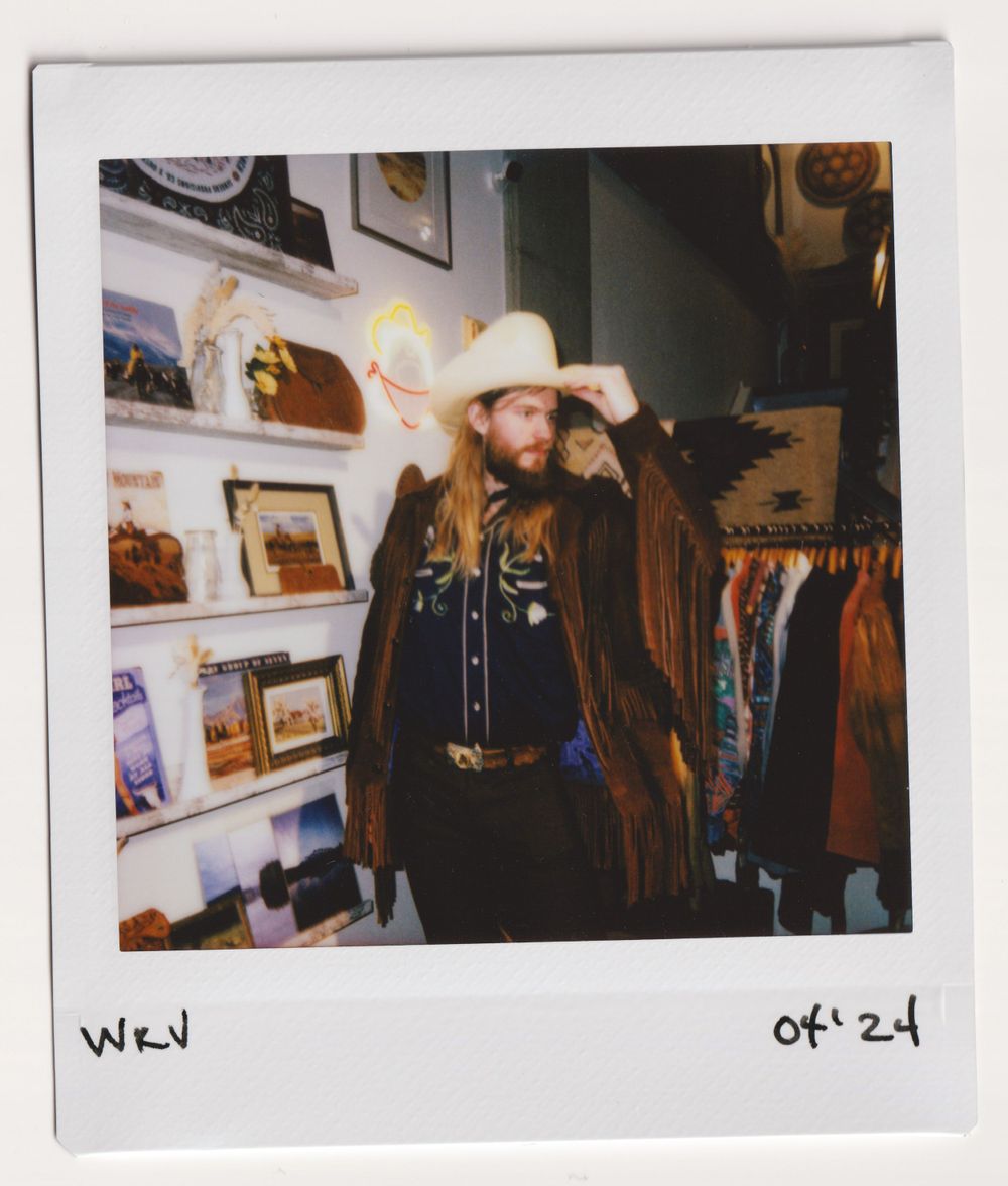 Person in black cowboy hat and western wear browsing vintage clothing store with wall displays.