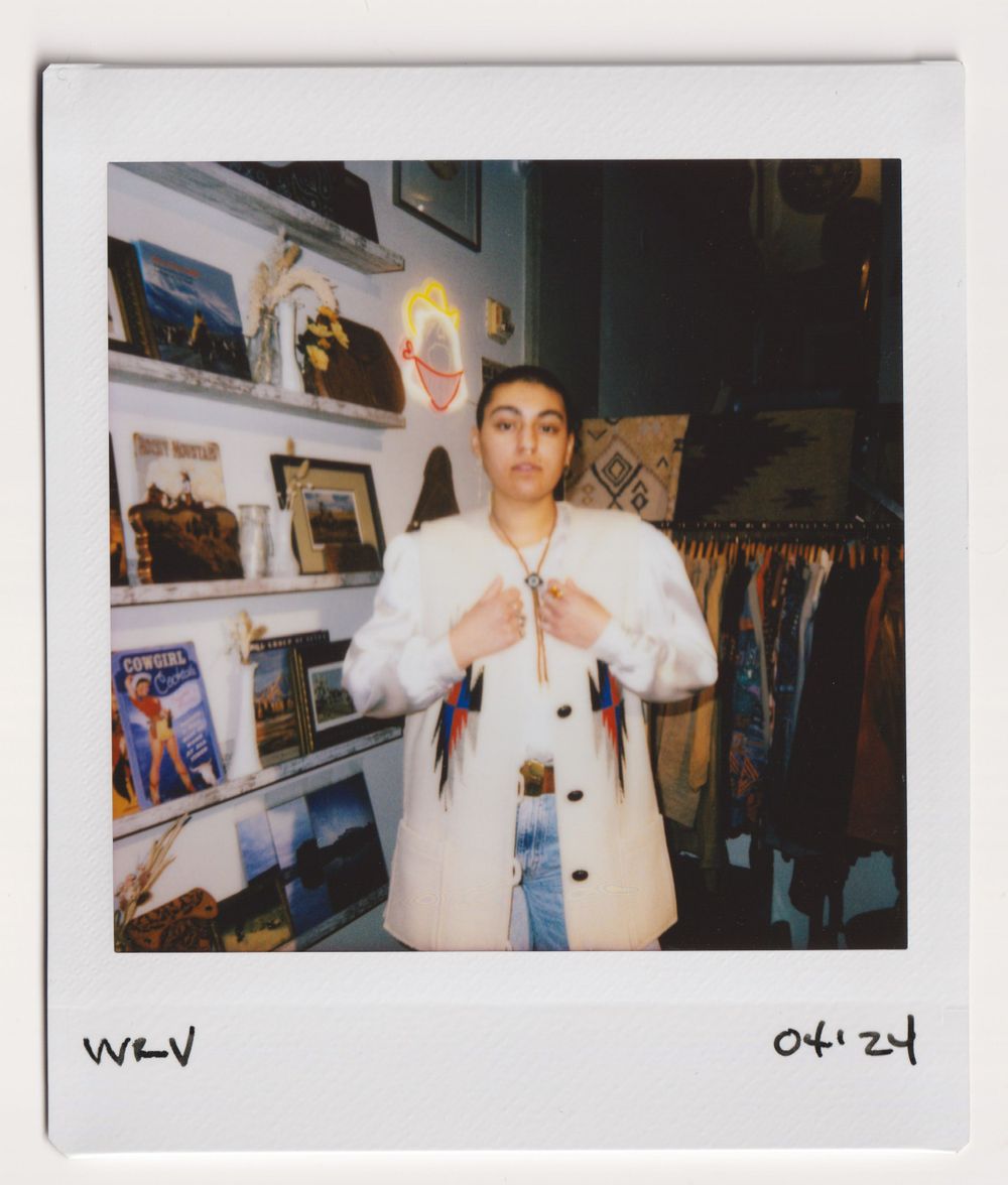 Shopper in white button-up shirt poses in front of wall display at vintage clothing store.