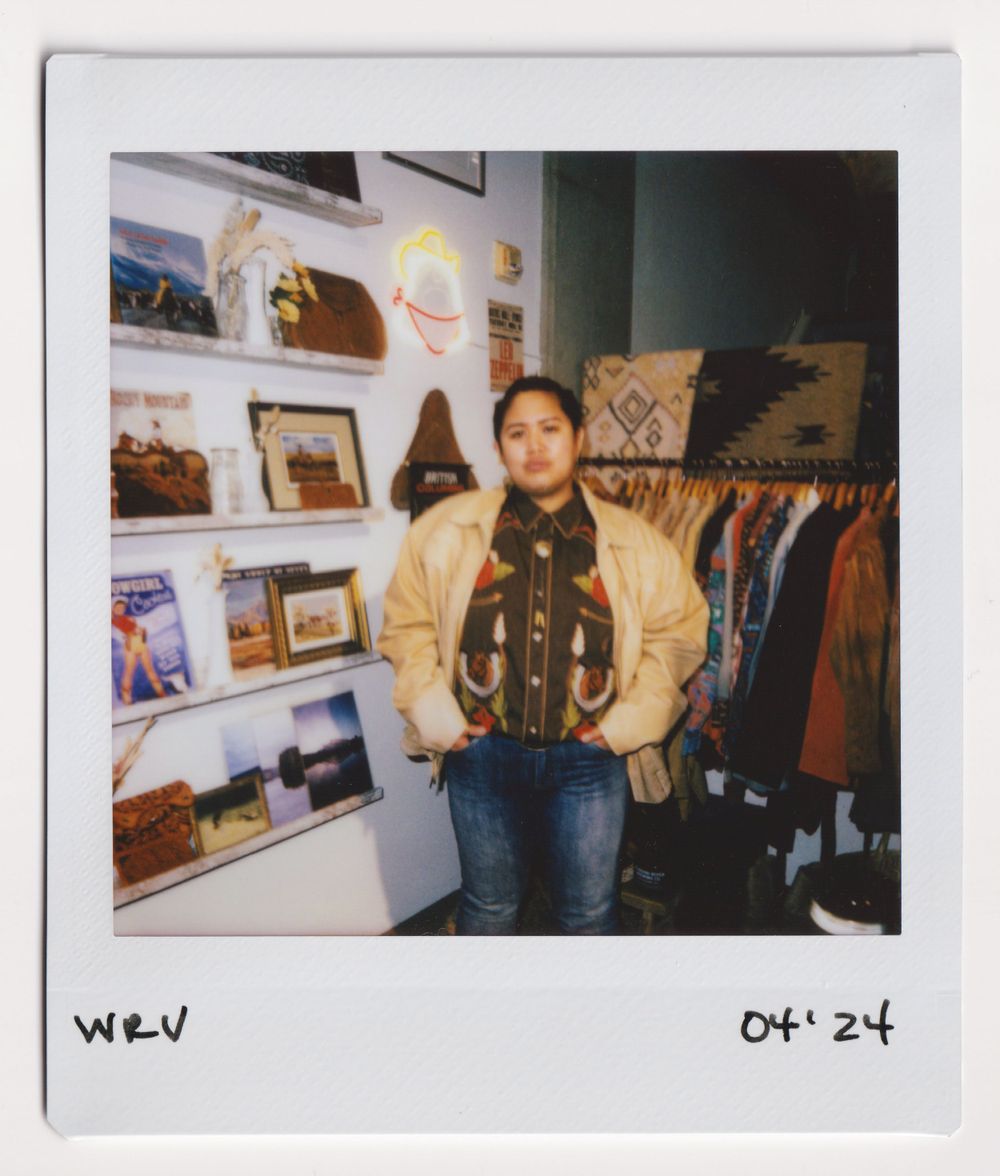Customer wearing tan jacket and scarf posing near clothing racks in vintage store.