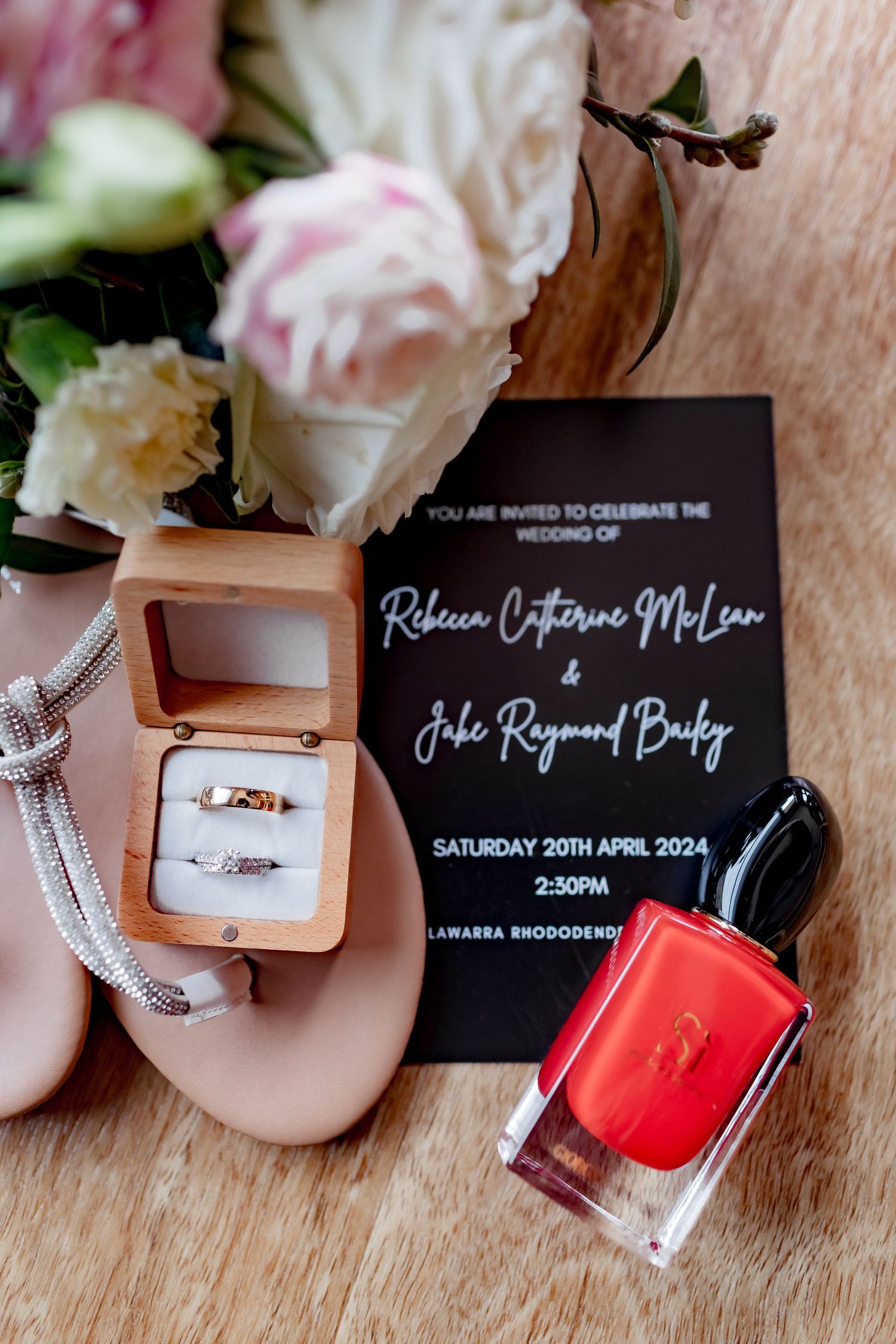 Wedding invitation with rings, pink flowers, and red nail polish arranged on wooden surface.