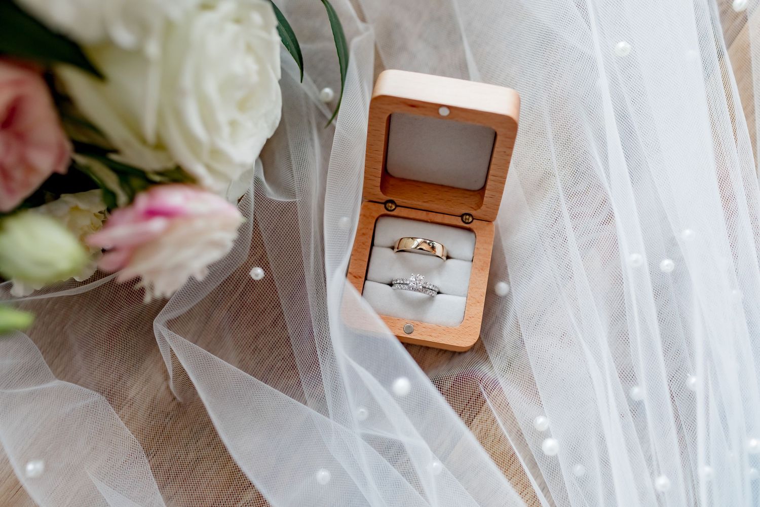 Wedding rings displayed in wooden box with white roses and veil in background.