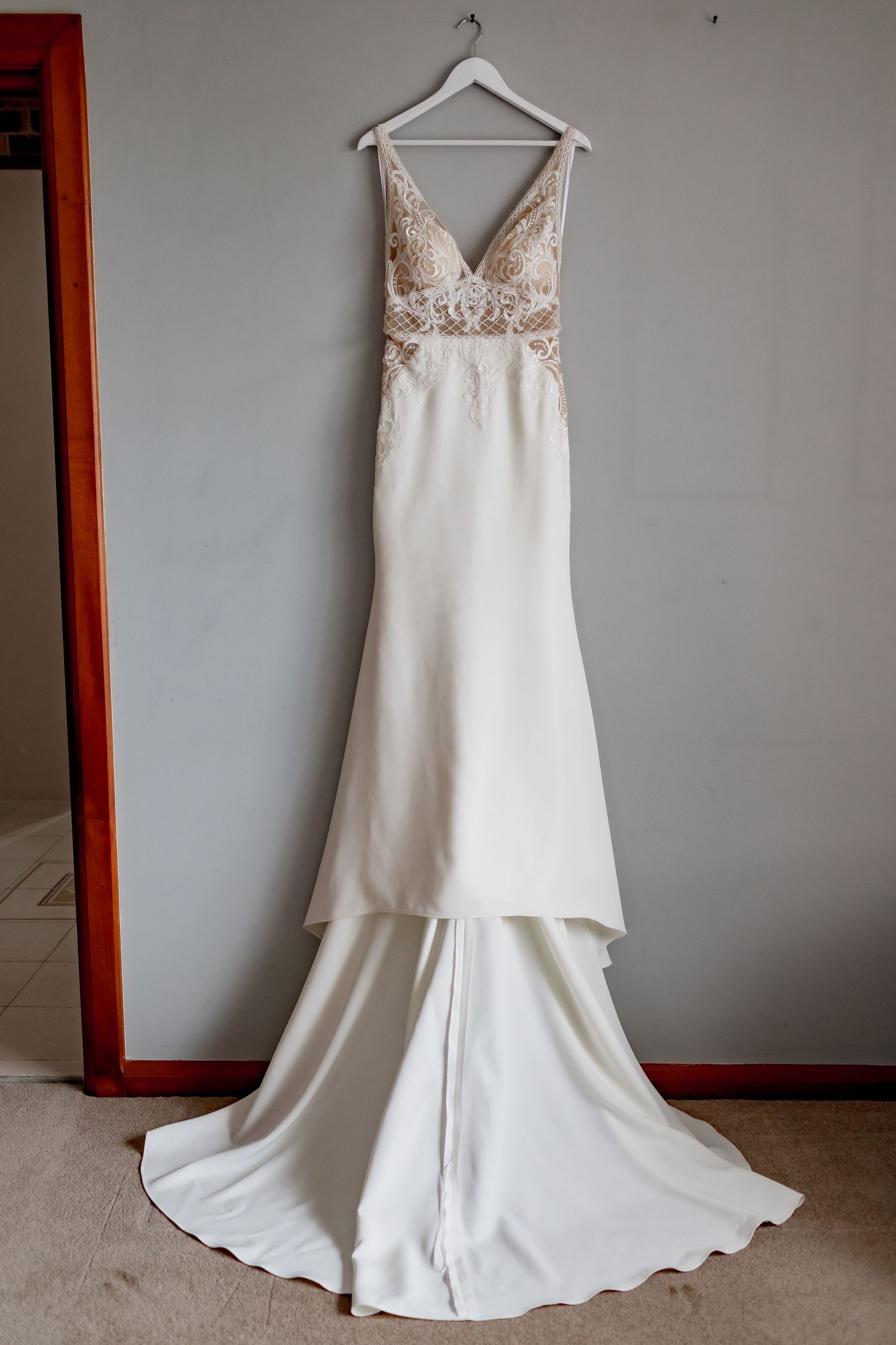 Elegant white wedding dress with lace details and plunging V-neckline hanging against grey wall.
