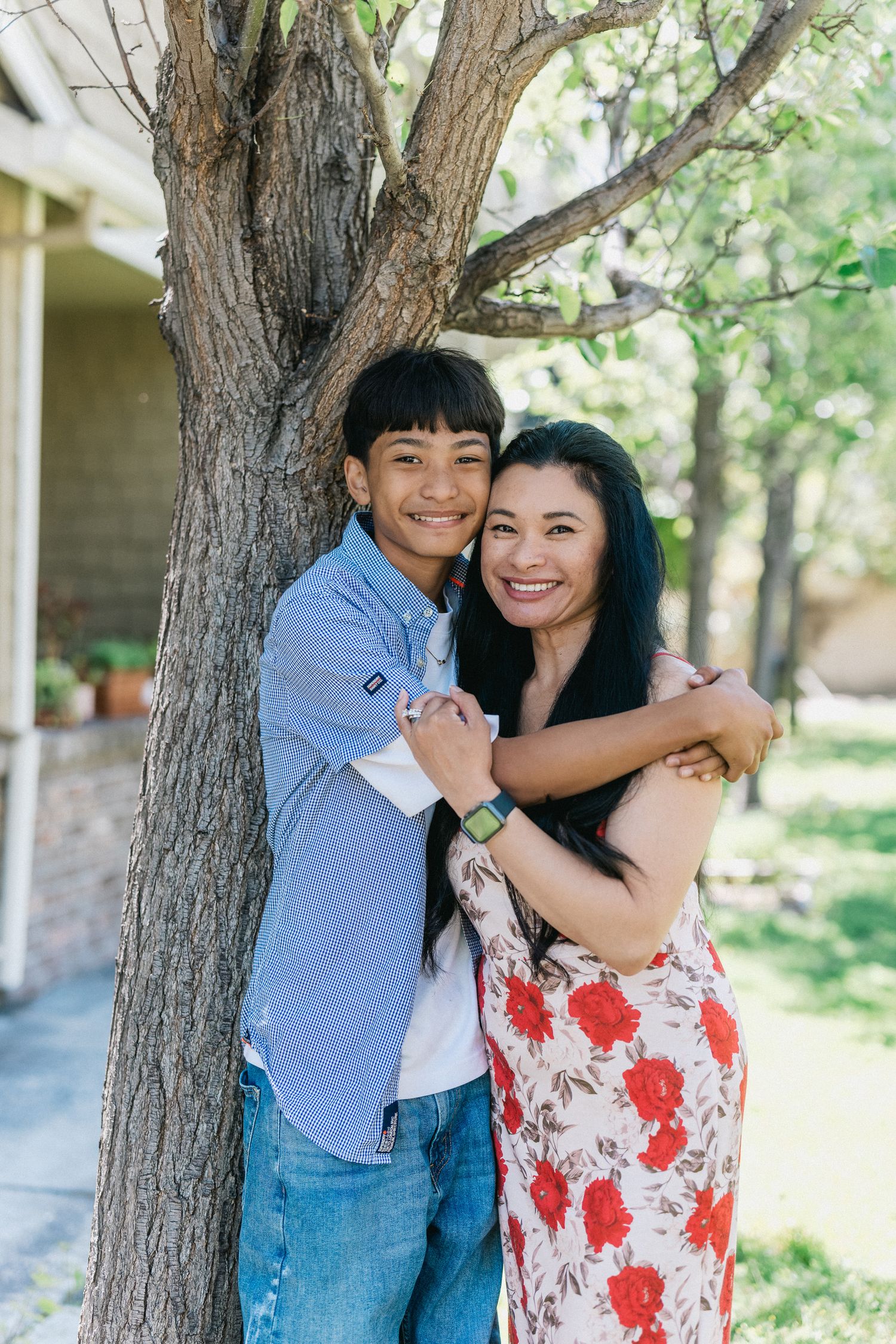 Couple embraces by a large tree trunk on a sunny day in front of a brick building.