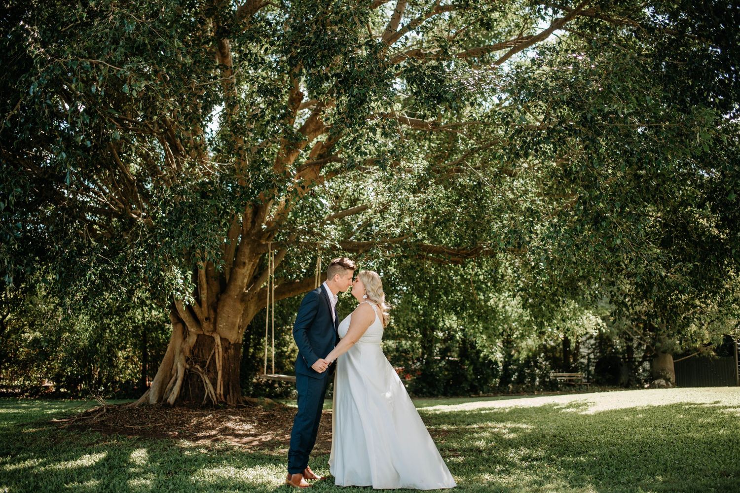 A couple shares a romantic moment on their wedding day under towering trees in a garden setting.