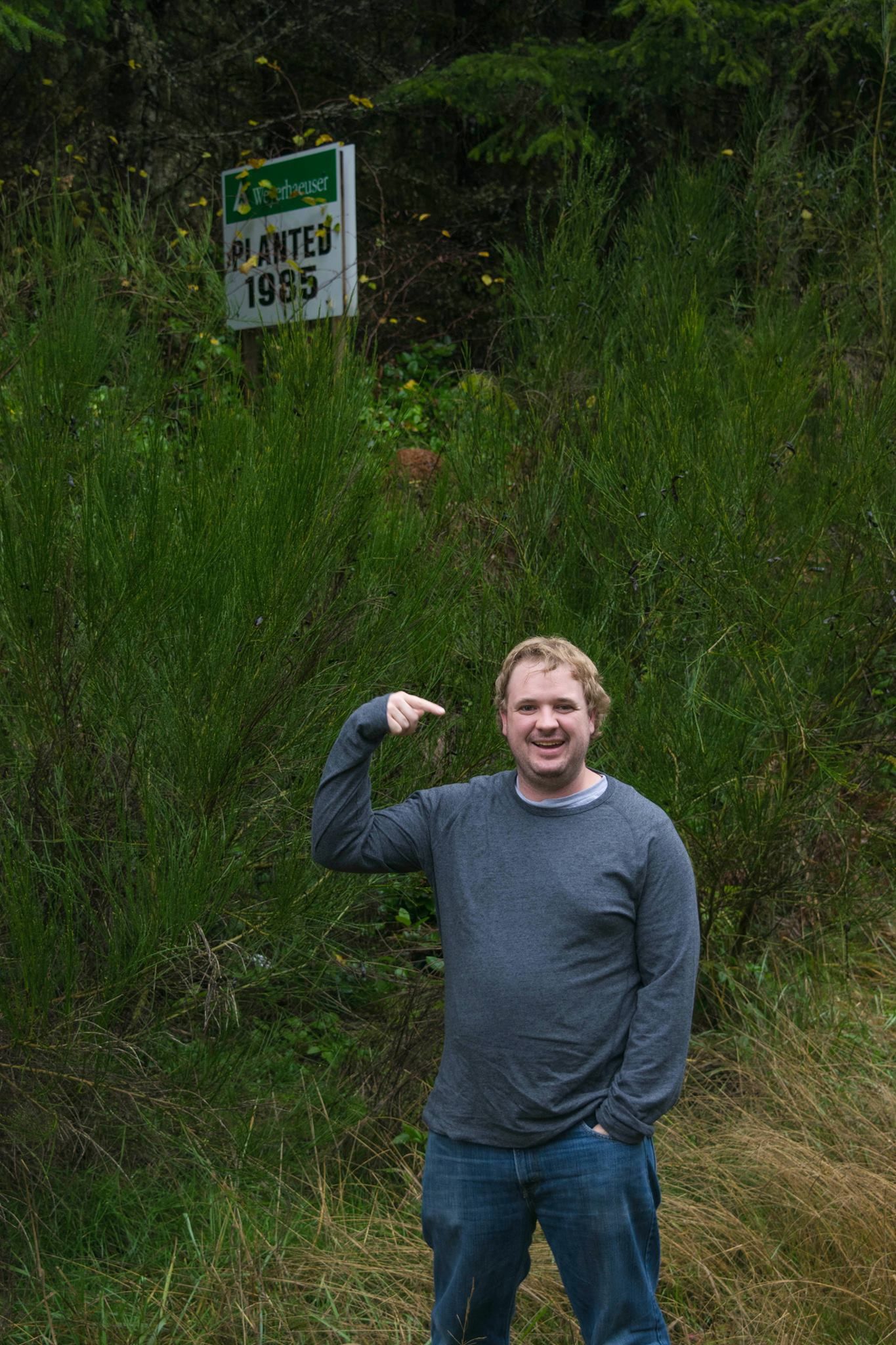 Visitor poses next to elevation marker sign surrounded by tall grass and greenery.