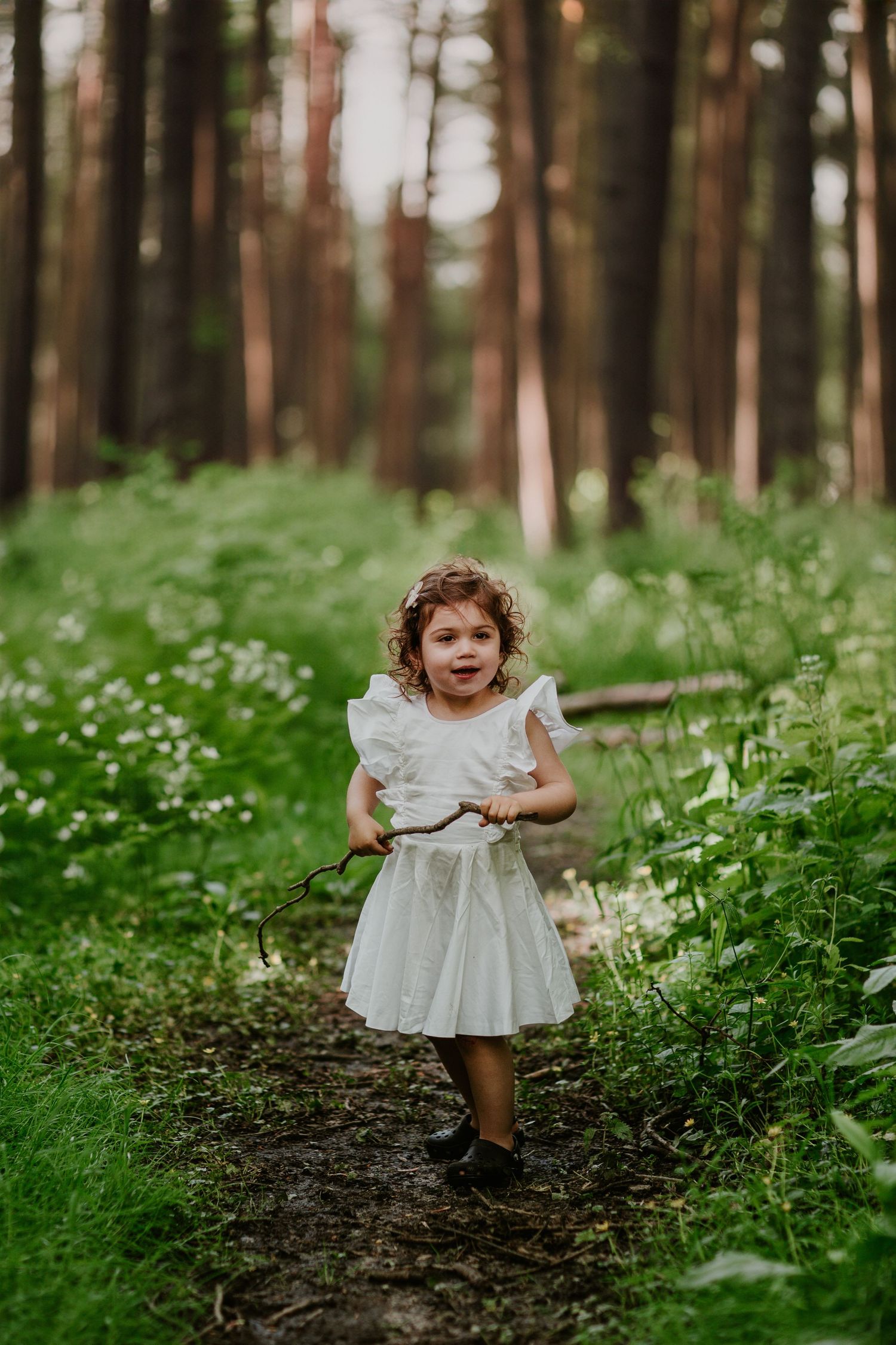 Child in white dress running along a woodland path surrounded by wildflowers and pine trees.