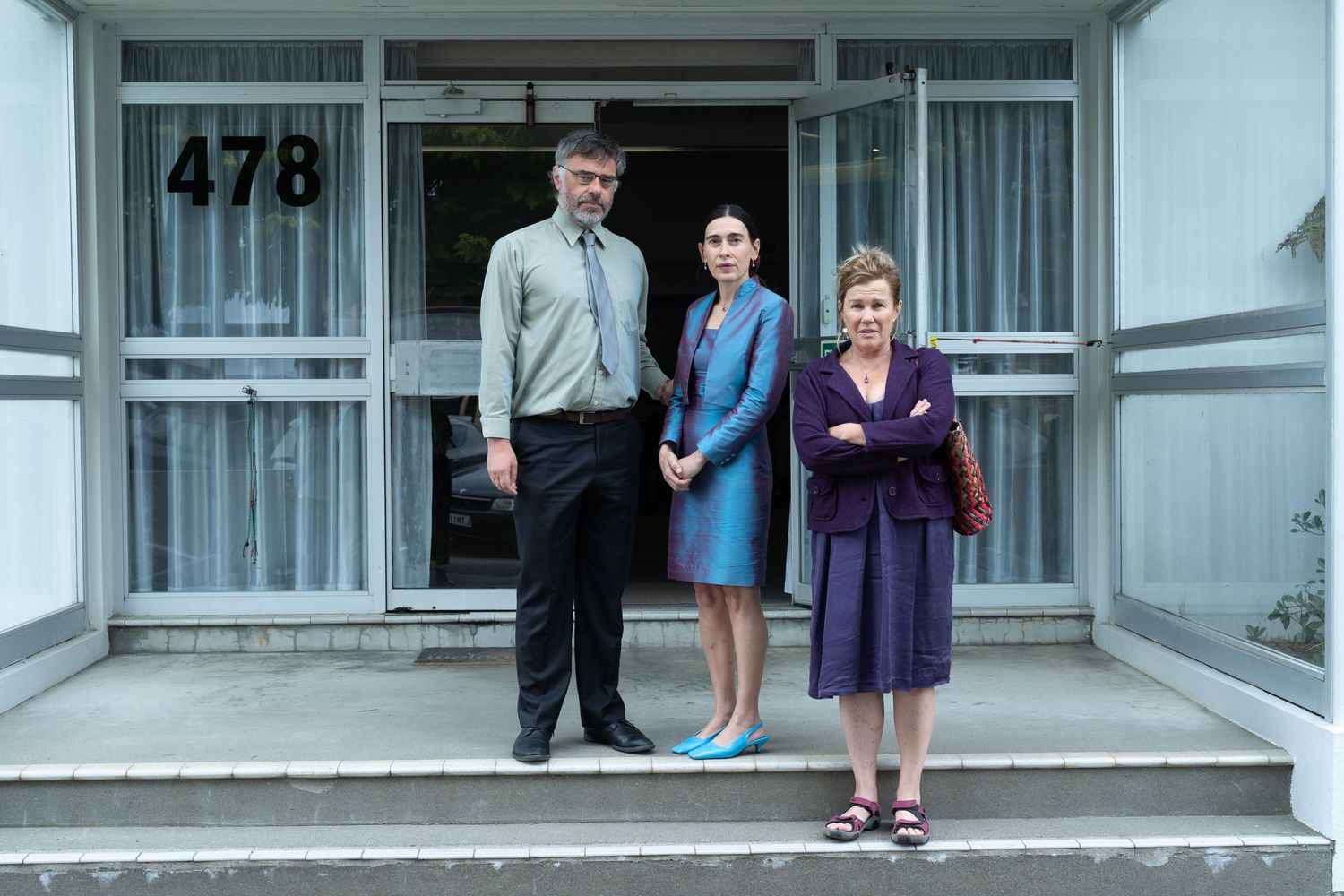 Jemaine Clement, Victoria Haralabidou and Robyn Malcolm standing in front of a building in character