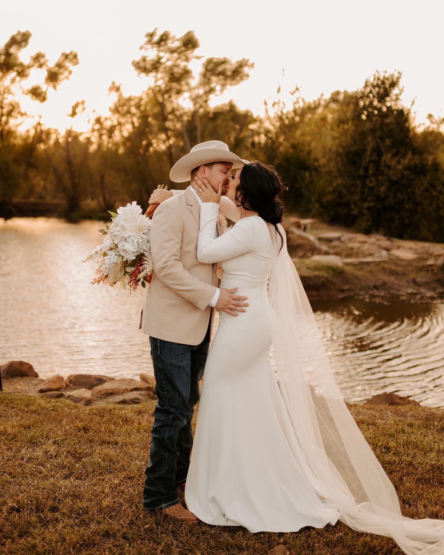 Romantic sunset wedding portrait by lake with couple in white dress and cowboy hat. Oklahoma Wedding Photographer