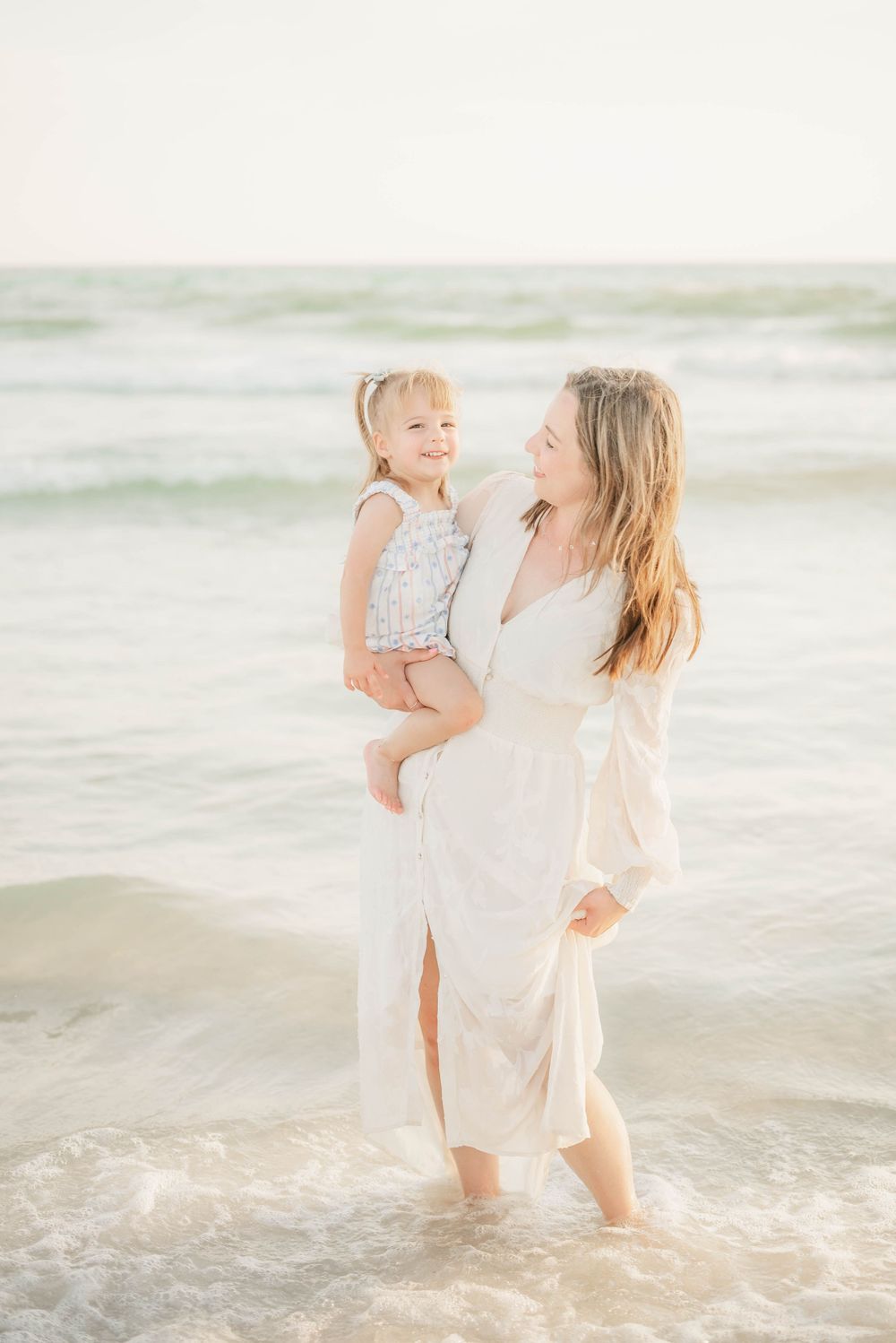 Mother and child embrace while standing in gentle ocean waves during golden hour.